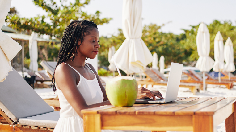 Pretty young woman sitting at table on sandy beach, enjoying fresh coconut cocktail and working on laptop.